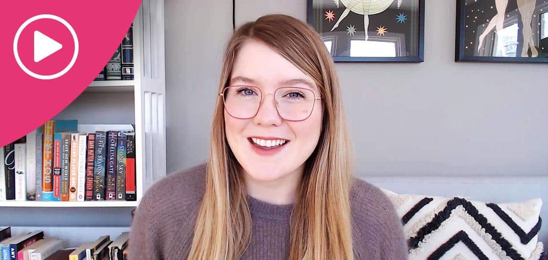 A person with long hair and glasses smiles at the camera. They are indoors, with shelves of books to one side and framed pictures on the wall. A pink play button icon is in the top left corner.