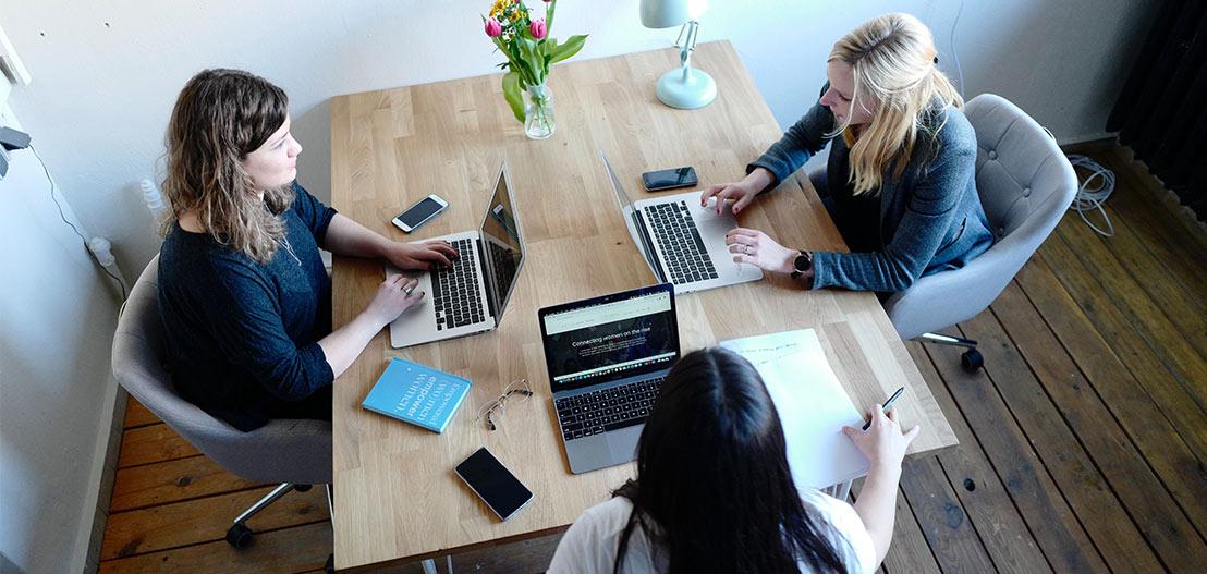 Three people sit at a wooden table with laptops, phones, and a notebook. A vase of flowers is in the center. They appear to be working or collaborating in a modern office space with natural lighting.