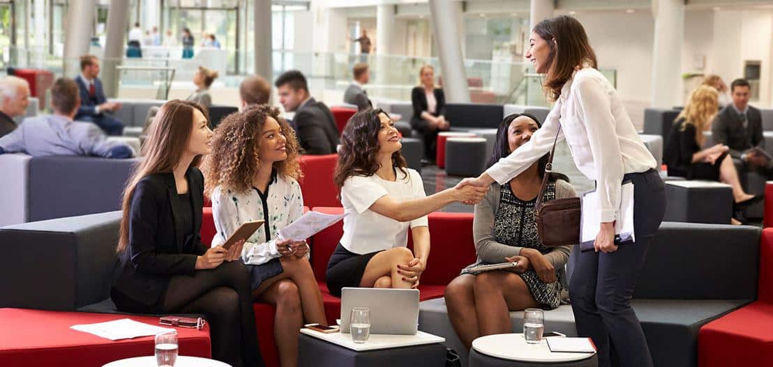 A group of four women are seated on modern couches in a bright office lounge, talking and holding documents. Another woman stands, smiling and shaking hands with one of them. People are gathered and conversing in the background.