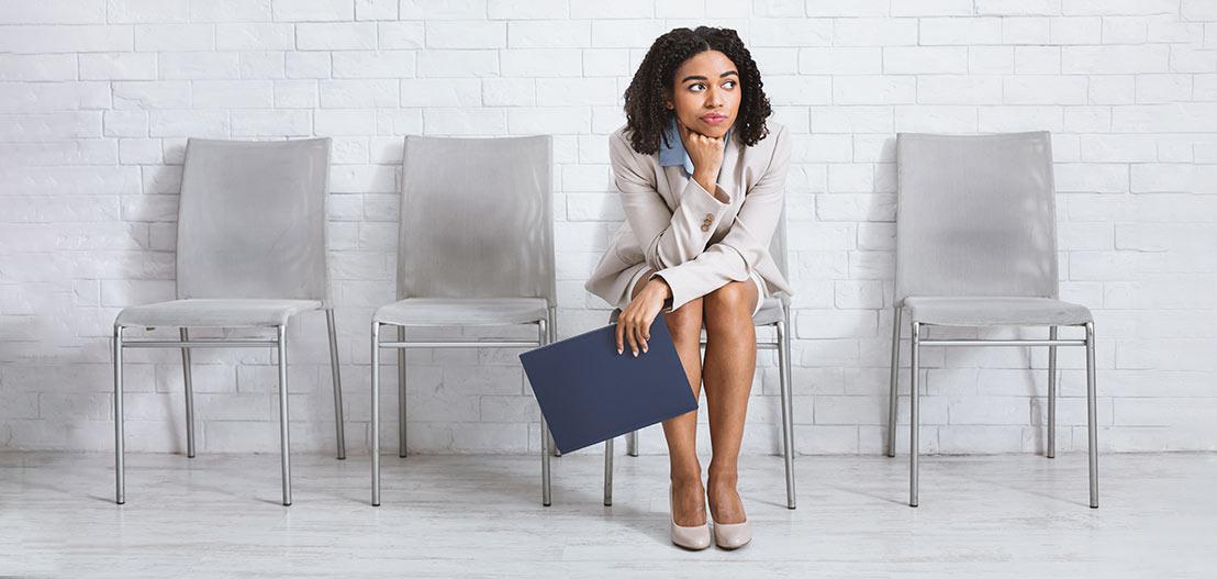 A woman in business attire sits on a chair against a white brick wall, holding a blue folder. She appears thoughtful, with three empty chairs beside her.