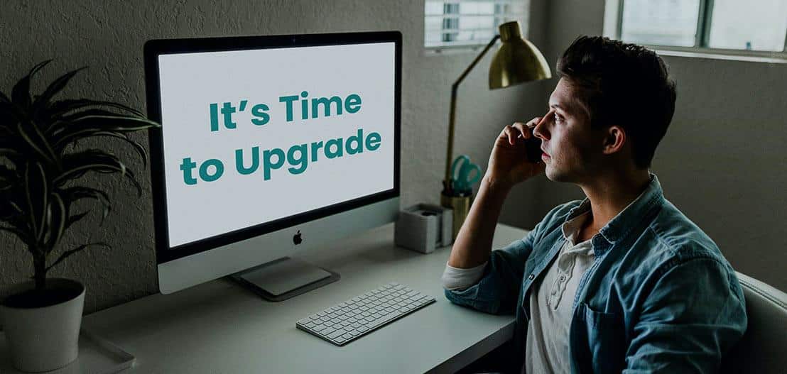 A person sits at a desk, immersed in a phone call while facing their computer. The screen boldly displays "It's Time to Upgrade Your Experience." A keyboard, mouse, and potted plant adorn the surface, with a lamp softly glowing in the background.