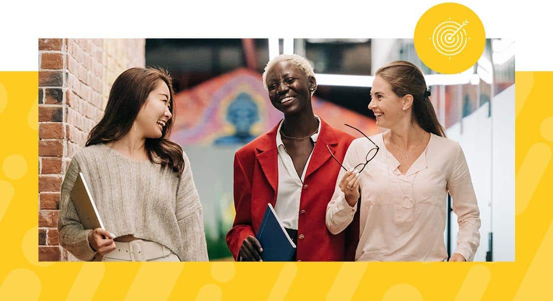 Three colleagues stroll through a well-lit hallway, their laughter echoing off the brick walls adorned with abstract art. One holds a tablet, another carries a blue folder and glasses, while the third in a red jacket discusses the benefits of a four-day work week.