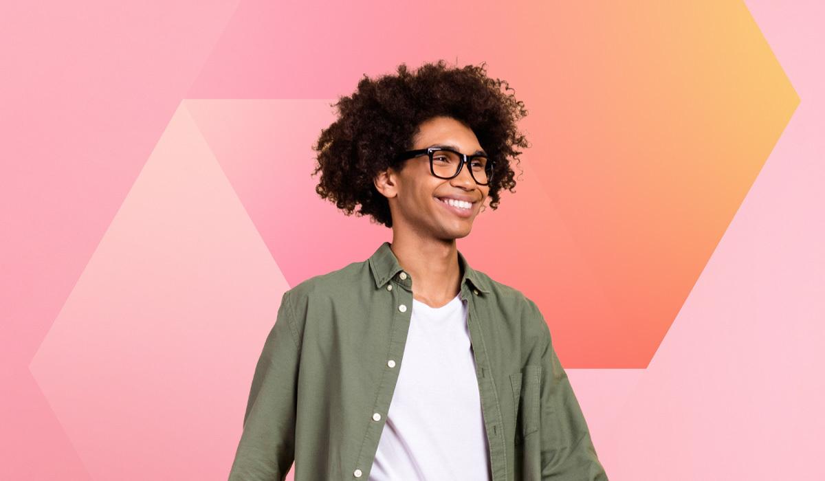 A smiling person with curly hair and glasses, donning a green shirt over a white T-shirt, stands confidently against a pink and orange geometric backdrop, reflecting the vibrant spirit often seen nurtured through employee development programs.