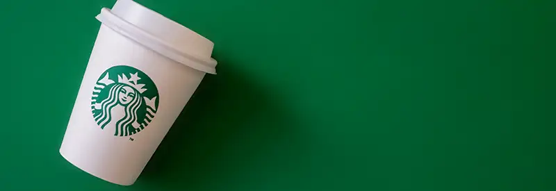 starbucks-banner A white Starbucks coffee cup with a lid and the green mermaid logo lies tilted on a solid green background, evoking a vibrant energy reminiscent of positive team dynamics.