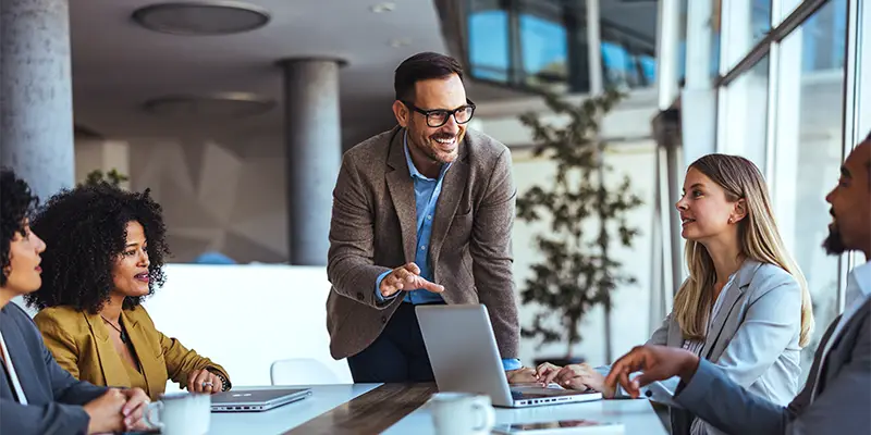 A group of four people sit at a conference table with laptops while a man in glasses and a blazer stands, smiling and speaking to them, fostering positive group dynamics in a bright, modern office space.
