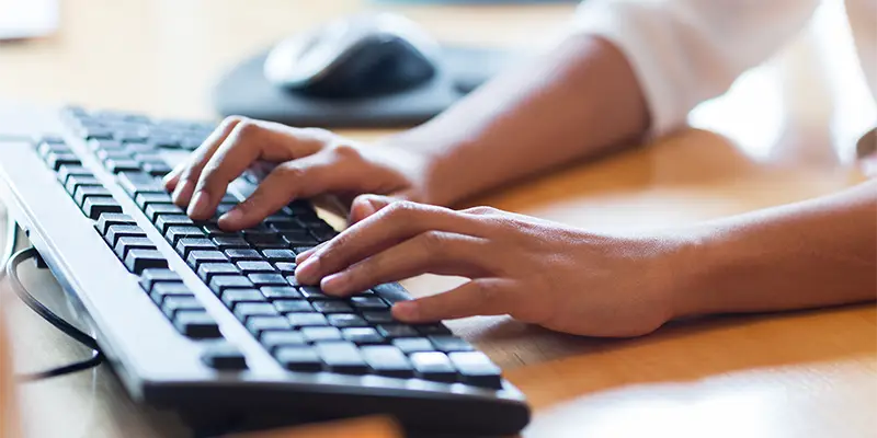 A person’s hands, demonstrating average typing speed, are typing on a black computer keyboard, with a computer mouse visible in the background on a wooden desk.