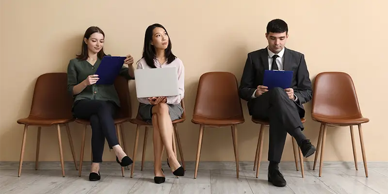 Three people sit on brown chairs against a beige wall, two with clipboards and one with a laptop on her lap, appearing to be part of a talent pool waiting for an interview.