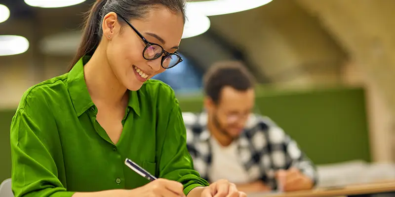 A woman in a bright green shirt and glasses smiles while writing at a desk in a classroom, exploring enneagram for hiring. A man in the background is also writing, with blurred lights and green walls around them.