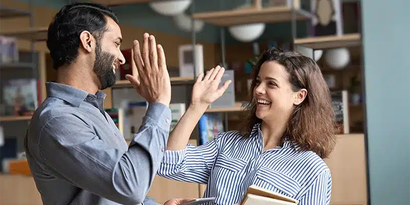 Two colleagues smiling and giving each other a high five in an office setting, highlighting positive teamwork and employee retention strategies. The woman is holding a notebook and a pen, with bookshelves and office supplies in the background.