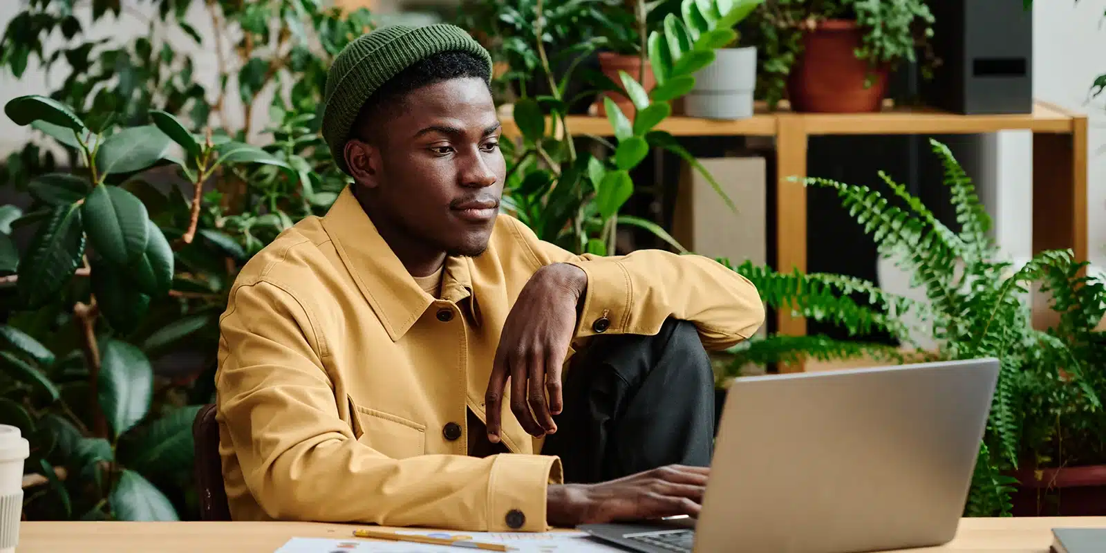 A person in a yellow jacket and green beanie sits at a desk with a laptop, surrounded by green plants, appearing focused on their work, embodying the Gen Z AI-driven approach to productivity.