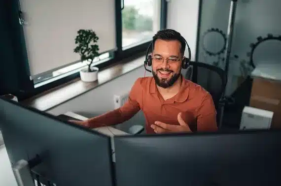 A bearded man with glasses and a headset sits at a desk with two computer monitors, smiling and gesturing as he talks.