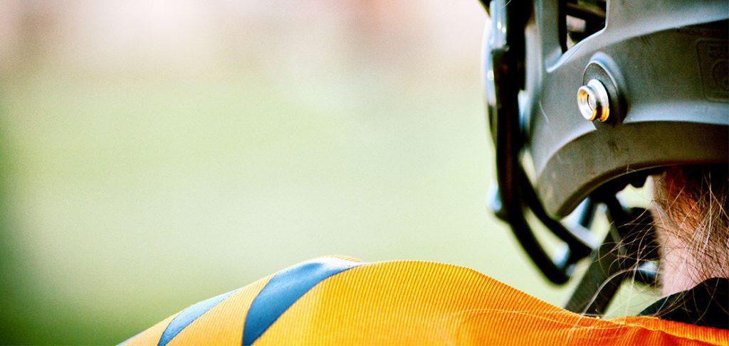 Close-up of a person wearing an American football helmet and jersey with black and orange stripes, viewed from behind. The blurred background emphasizes the focus on the helmet and jersey, symbolizing the dedication to finding work-life balance through sport.