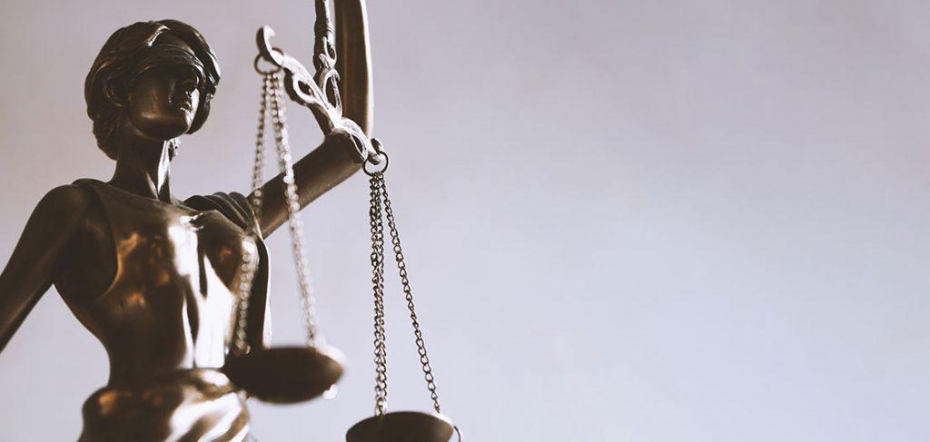 Bronze statue of Lady Justice holding balanced scales and a sword. The figure is blindfolded, symbolizing impartiality. The background is plain and softly lit, drawing focus on the statue's details and symbolism.