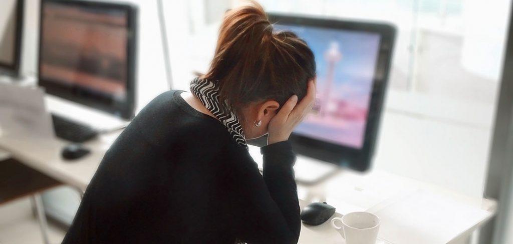 A person with a ponytail, wearing a black and white outfit, is sitting at a desk, holding their head with one hand. A white mug is on the desk. Computer screens showing blurred content are in the background.
