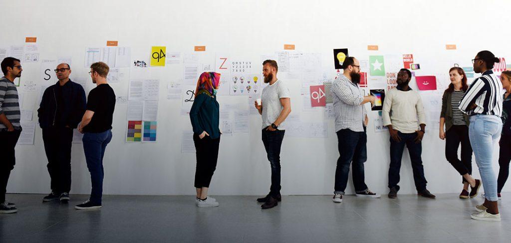 Nine people stand and chat in front of a large white wall covered with various colorful posters and papers. They appear to be in a casual discussion, possibly a creative meeting or workshop, in a bright, minimalistic room.