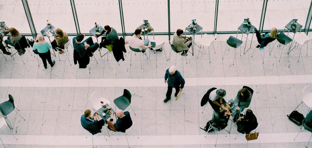 Aerial view of people sitting and talking at small tables in a modern, bright atrium. The glass wall allows natural light in. Some are on phones, others engaged in conversation. A person walks through the space. Minimalist design with gray flooring.