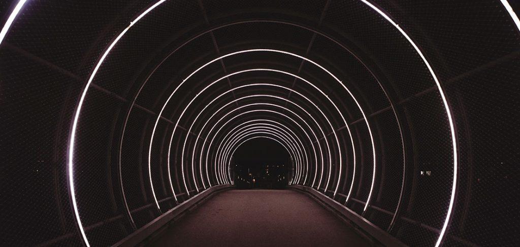 A tunnel with glowing white concentric circles illuminates the path. The rings create a futuristic and symmetrical effect against the dark, enclosed background. The ground and distant lights are faintly visible.