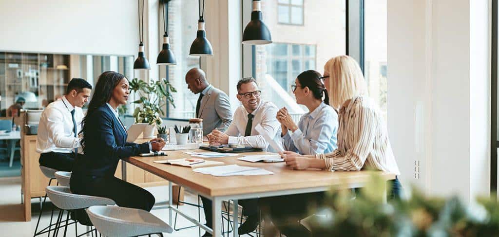 A diverse group of six people are engaged in a meeting around a large wooden table in a modern office setting. Natural light streams through large windows, and the space is decorated with hanging lights and plants. Papers and devices are on the table.