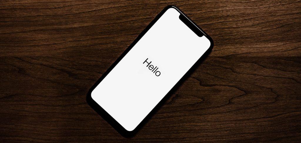 A smartphone with a black frame displays the word "Hello" on a white screen. The phone is laying on a wooden surface, slightly angled to the left.