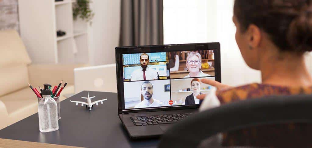 A person is participating in a video call on a laptop. The screen displays four participants. The desk has a water bottle with pens, a small airplane model, and a blurred living room background is visible.
