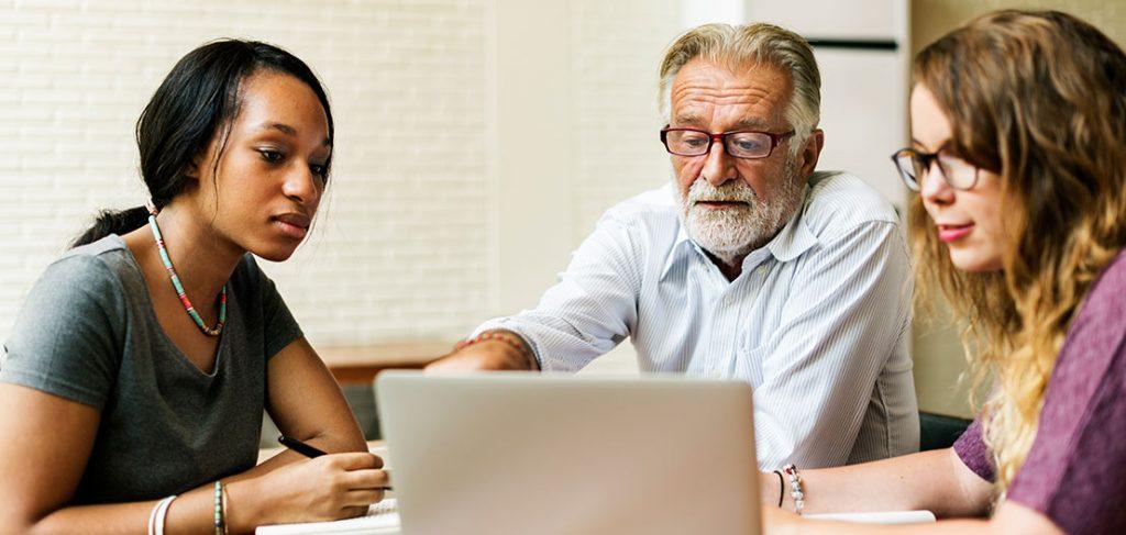 A diverse group of three people are seated at a table, engaged in a discussion while looking at a laptop. An older man with glasses points at the screen, while two younger women listen attentively, taking notes.