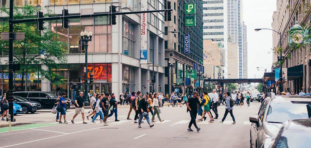 A busy city intersection with many pedestrians crossing the street. Tall buildings with various signs and storefronts line the street. Cars are stopped, and the scene is bustling with activity on a clear day.