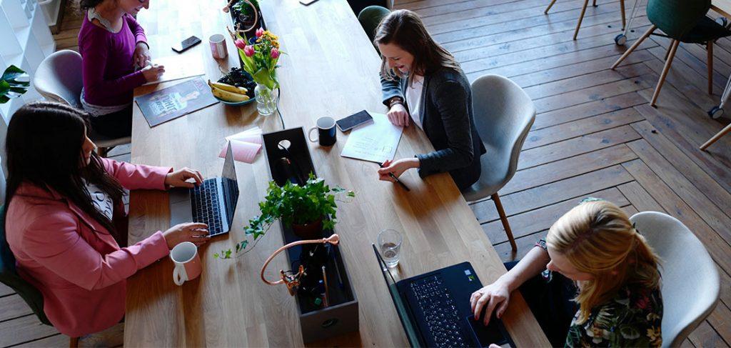 Four people are working at a long wooden table with laptops, notebooks, and coffee cups. The table is decorated with plants and a vase of flowers, and the room has wooden floors and a casual atmosphere.
