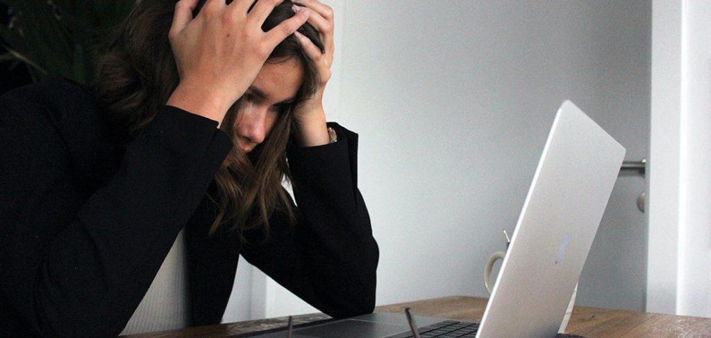 A person sits at a wooden table, looking stressed while holding their head in their hands. An open laptop is in front of them. Glasses lie on the table next to the laptop. A mug is partially visible in the background.