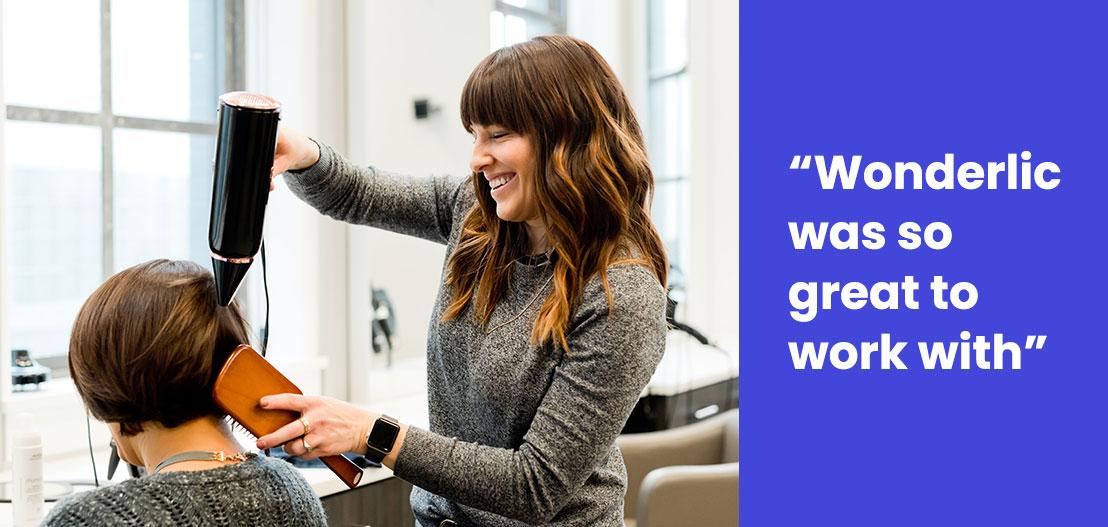 A stylist blow-dries a client's hair in a salon, both smiling. The stylist, with long brown hair and wearing a gray sweater, exemplifies the seamless gain of skills through hands-on education. A blue panel on the right reads: "Wonderlic was so great to work with.