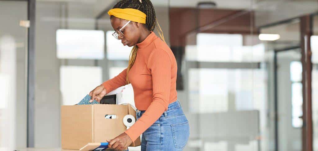 A person in an orange turtleneck and yellow headband is packing items into a cardboard box. They are in a bright office setting with large windows.