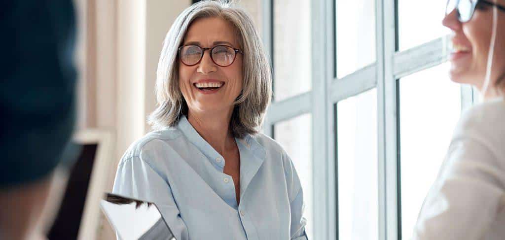 A smiling woman with gray hair and glasses, wearing a light blue shirt, sits in a bright room by a window. She appears engaged in conversation with two other people who are partially visible.