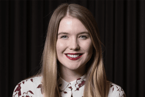 A person with long blonde hair is smiling at the camera, as if preparing for a hiring assessment. They are wearing a white shirt with a maroon leaf pattern. The background is dark with vertical lines, adding an air of mystery to the scene.