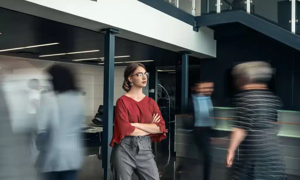 A woman in a red blouse and gray pants stands confidently with arms crossed in a modern office building, highlighting her strong role amid the fast-paced team dynamics as people around her blur by, suggesting movement and busyness.