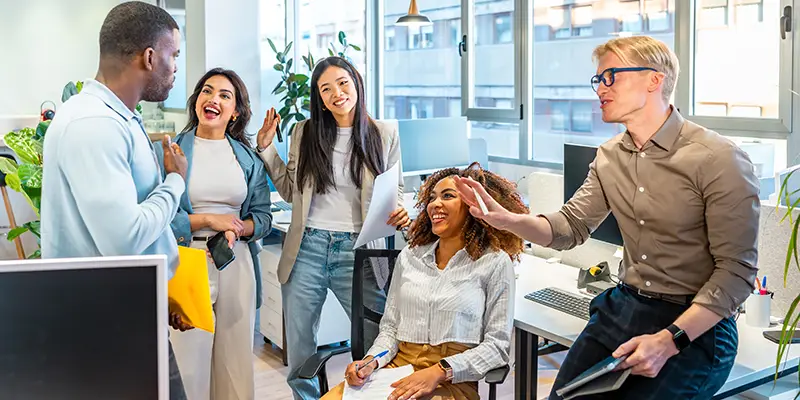 Five colleagues in a modern office laugh and interact energetically. One stands with a folder, while another offers a high-five to a seated coworker taking notes. The mood is lively and positive.