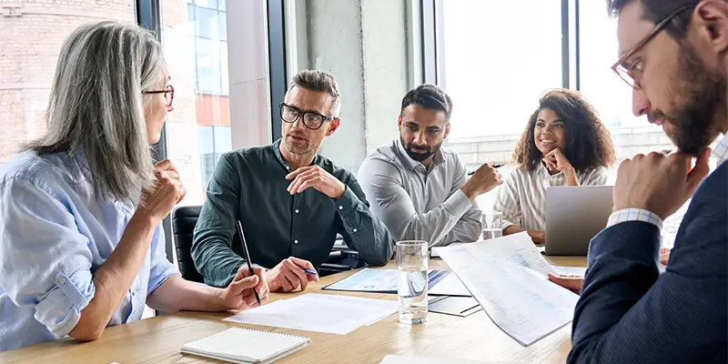 Five people sit around a table in a bright office, discussing team roles with papers, a notebook, and a laptop in front of them. One woman speaks while others listen attentively.