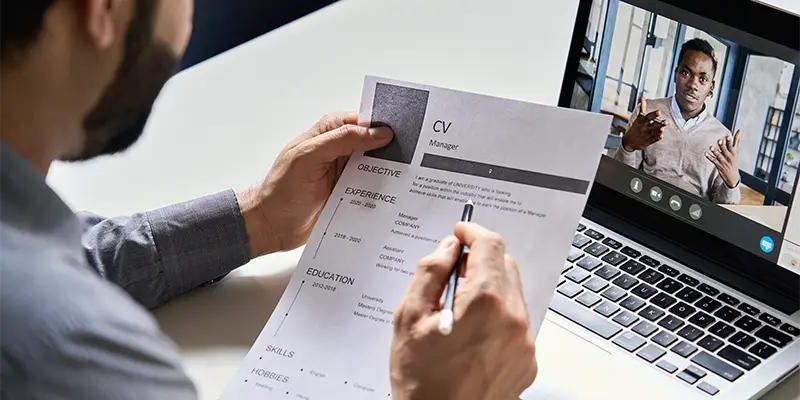 A person holds a resume and a pen while attending a video interview on a laptop, where another individual is speaking on the screen in a modern office setting.