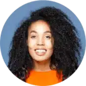 Smiling person with long, curly black hair wearing an orange top, photographed against a plain blue background.