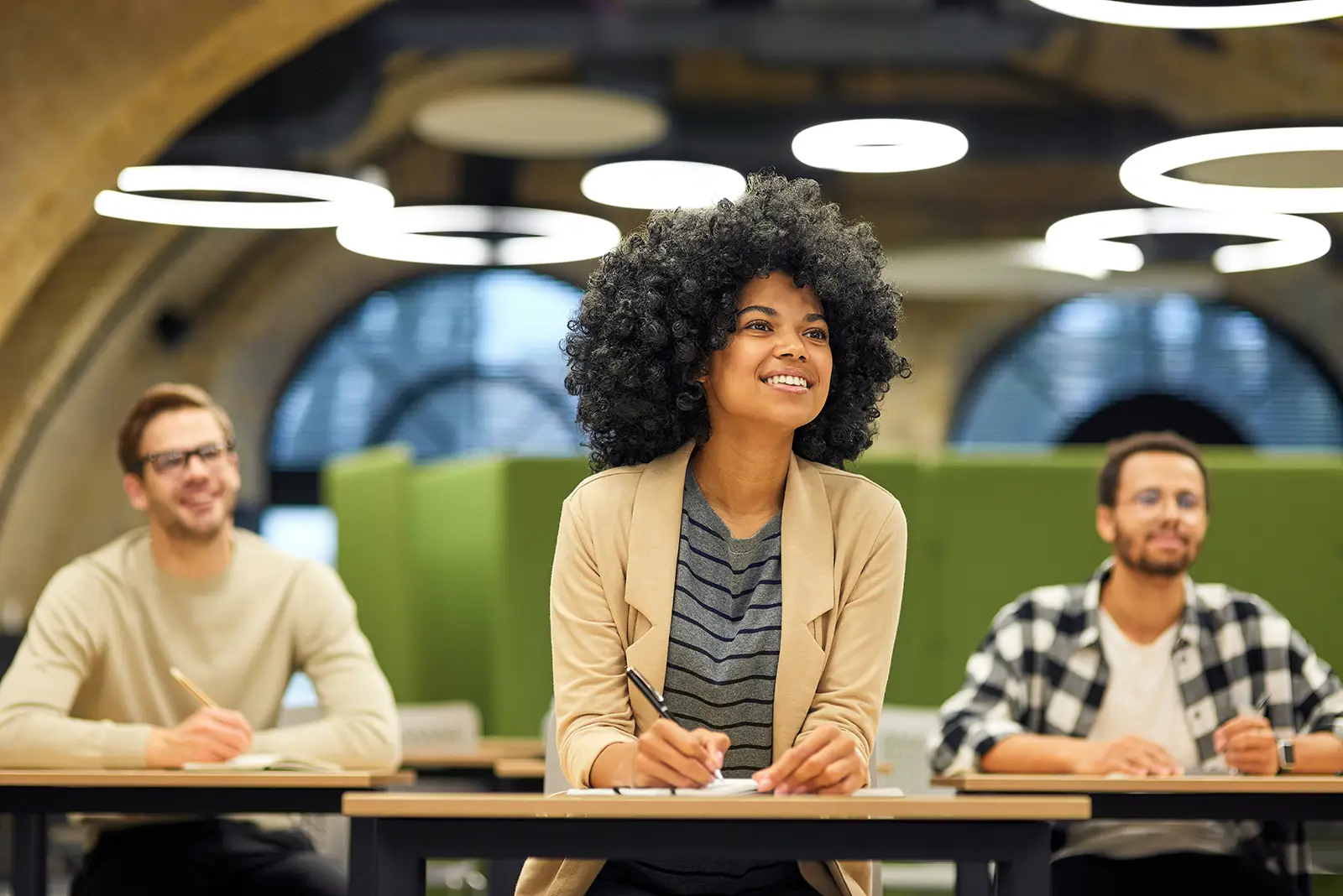 Three people sit at desks in a modern classroom. A woman in the center, smiling, writes in a notebook while preparing for Wonderlic certification. Two men on either side are also engaged, with blurred green partitions and circular lights behind them.