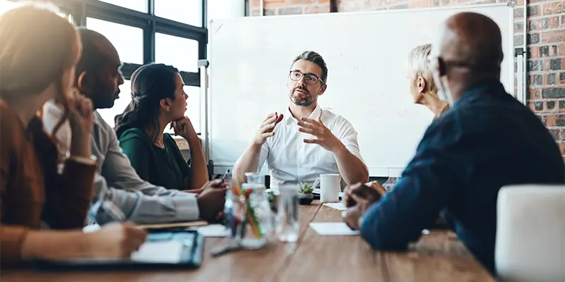 A group of five people sit around a table in an office, attentively listening to a man speaking about the enneagram in the workplace as he gestures with his hands in front of a whiteboard.
