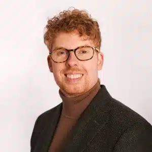 A smiling person with curly reddish hair and glasses, wearing a brown turtleneck and a dark blazer, poses in front of a plain light background.