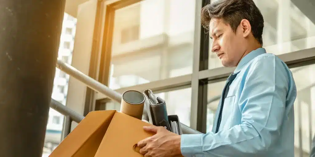 A man in a light blue shirt is holding a cardboard box filled with office supplies, including a rolled-up document, a mug, and a folder, reflecting changes in team dynamics as he stands near large windows in an office building.