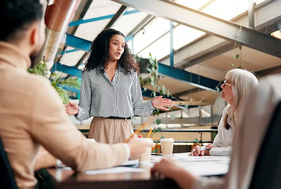 A woman stands and speaks to colleagues seated around a table in a modern office, gesturing with her hands as they discuss wonderlic certification. Papers, notebooks, and coffee cups are on the table, with sunlight streaming through large windows above.