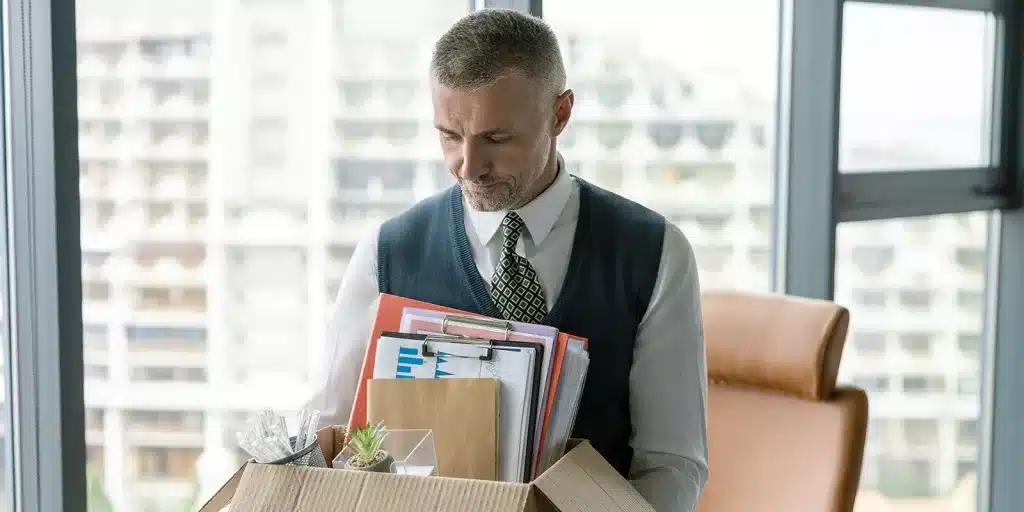 A man in business attire looks down at a box filled with office supplies and personal items, reflecting the reality of employee turnover as he stands in an office with large windows showing a cityscape outside.