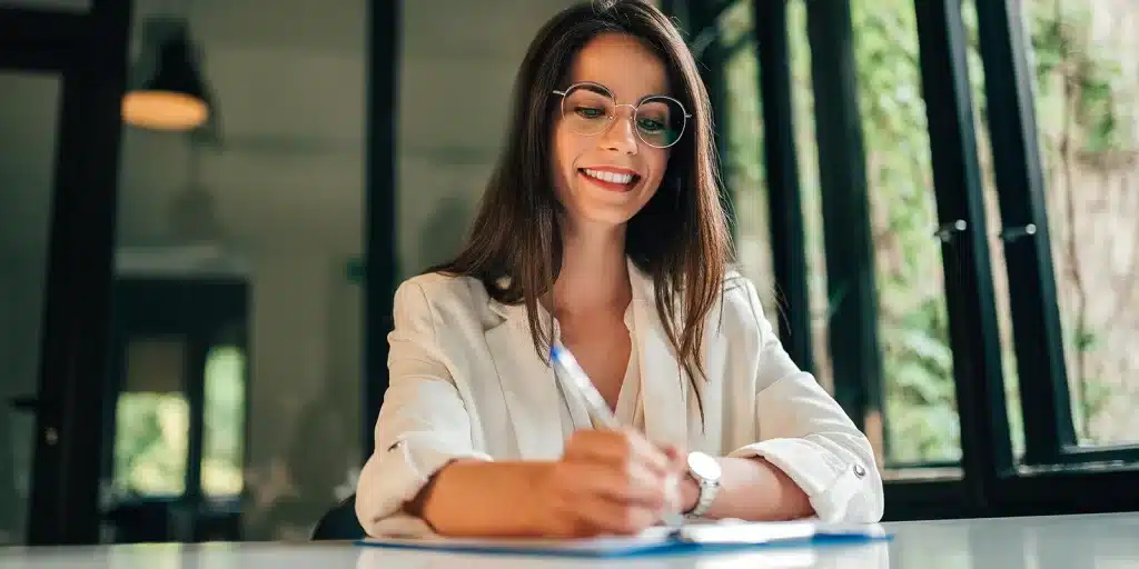 A woman with long brown hair and glasses smiles while writing on paper at a desk, wearing a white blazer. She appears to be completing an employee engagement survey as she sits in a bright room with large windows and greenery outside.