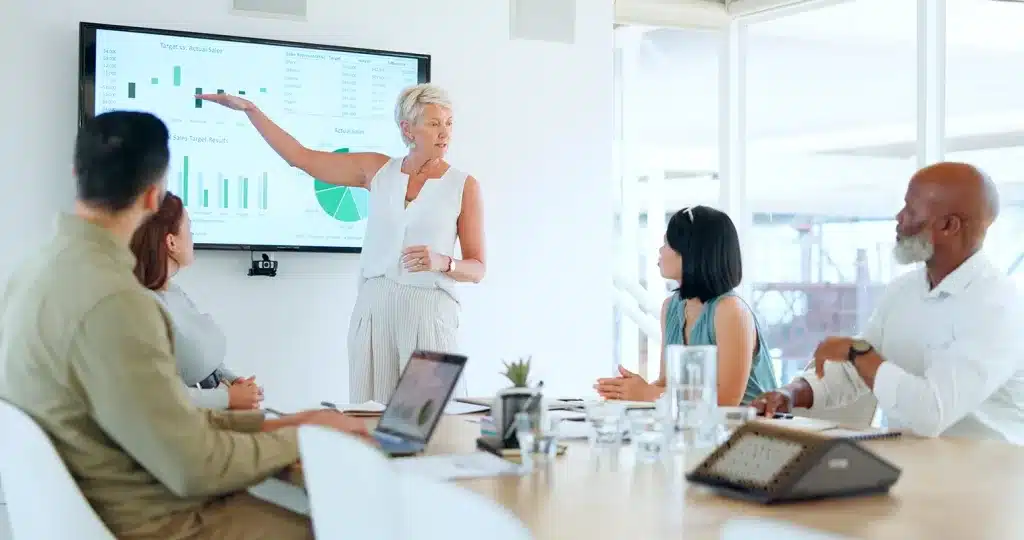 A woman stands and points to a presentation slide with charts on a screen, highlighting employee turnover rate benchmarks, while four colleagues sit around a conference table listening attentively.