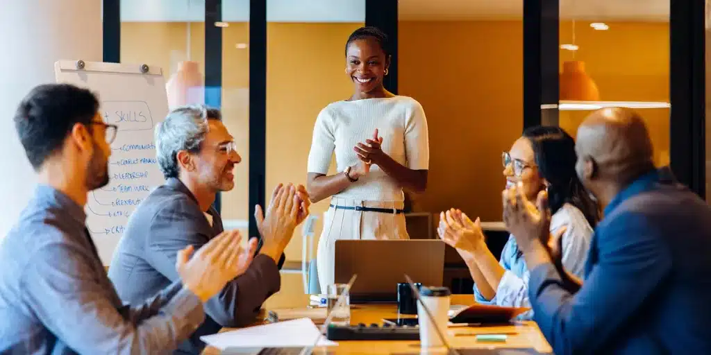 A group of five people sit around a table in an office, clapping and smiling at a woman standing and applauding. Their enthusiasm reflects strong employee engagement, while a whiteboard, laptops, and coffee cups are on the table.