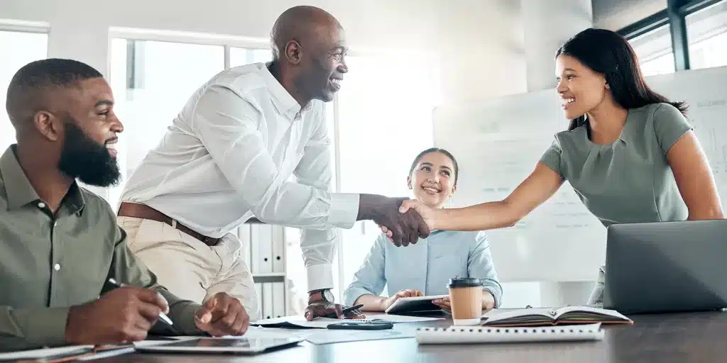 Four people in a modern office, two standing and shaking hands while two others sit, smiling. Laptops, notebooks, and coffee cups suggest a positive employee onboarding meeting.