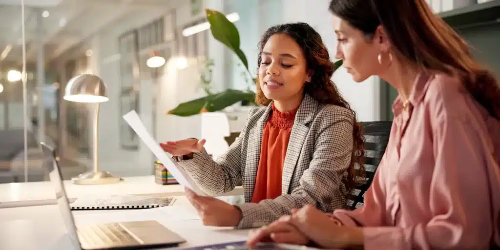 Two professional women sit at a desk reviewing a document together.